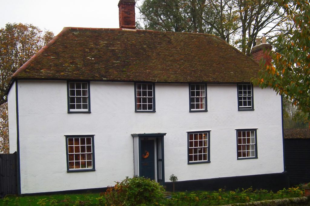 A white cottage with a dark tiled roof, a red brick chimney and tidy hedges